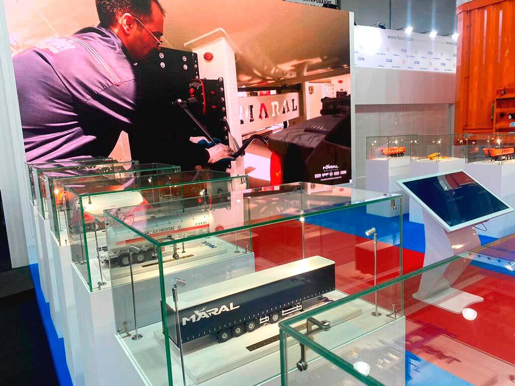 На фотографии представлен макет оранжевого полуприцепа с 12 колесами, выполненный с высокой детализацией. Полуприцеп имеет аккуратные линии и реалистичную отделку, что подчеркивает его предназначение для выставки. Фон изображения ненавязчивый, что позволяет сосредоточиться на модели техники.