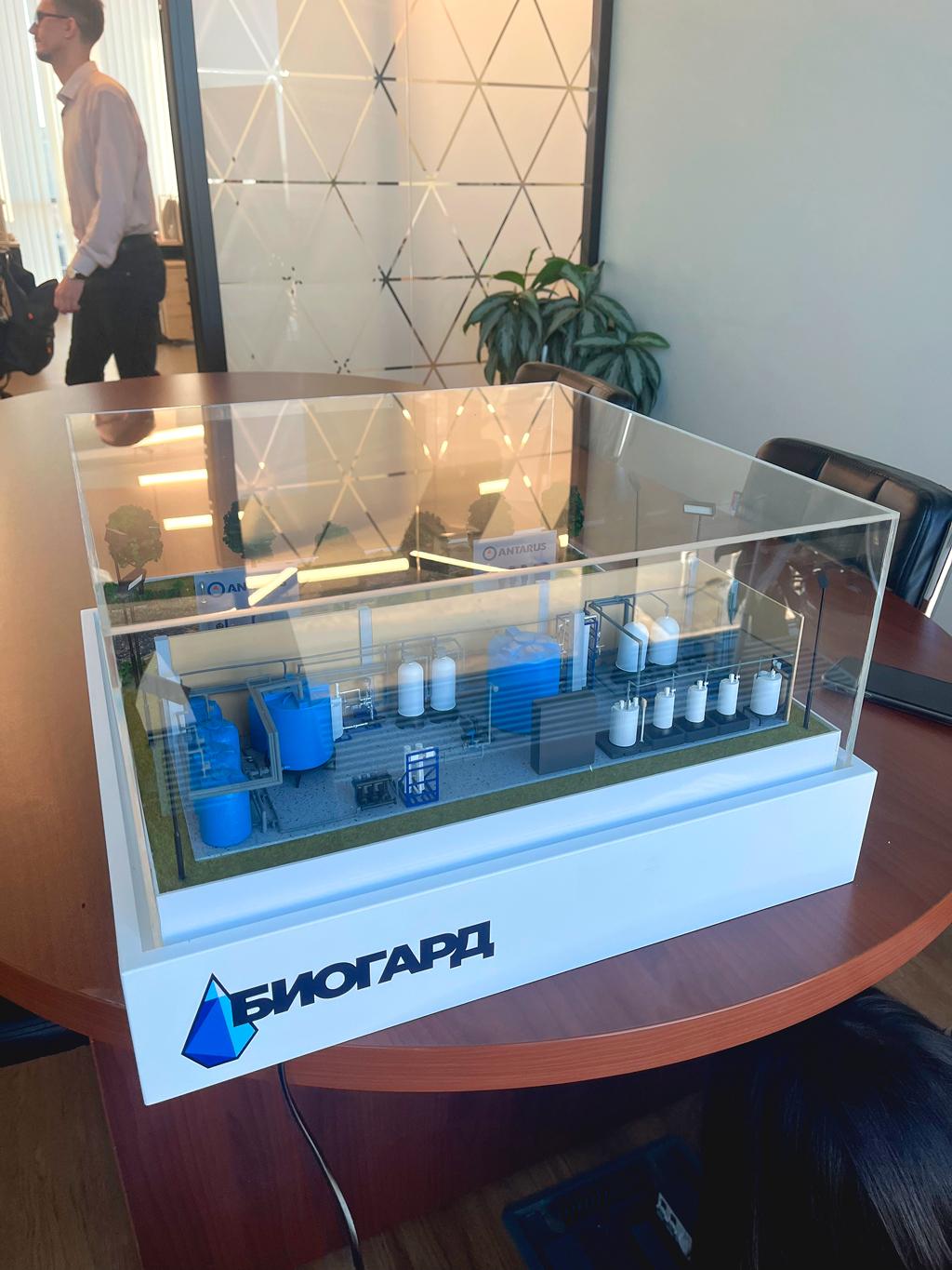 Макет водоподготовки и очистки водопроводной воды, заключенный в прозрачный акриловый бокс. Бокс стоит на белой подставке, на которой крупными синими буквами написано "БИОГАРД". Модель включает в себя миниатюрные резервуары (синие и белые), трубы и другие элементы промышленной инфраструктуры. Изображение создает впечатление современного, технологичного и профессионального окружения. Макет предназначен для демонстрации или презентации системы очистки воды.