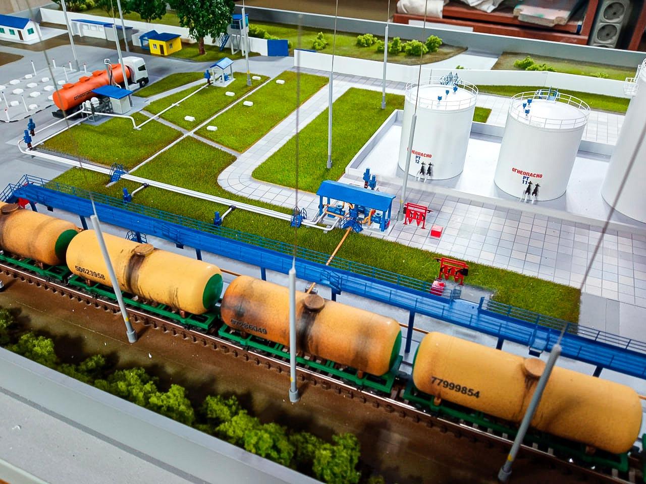  3D-масштабный макет перевалочного комплекса нефтепродуктов: показаны железнодорожные пути с составом цистерн для перевозки нефти, резервуарный парк с вертикальными стальными резервуарами, площадка для автоналива с моделями автоцистерн, элементы инфраструктуры (мачты освещения, дорожки, зоны озеленения). Демонстрирует технологическую схему хранения и отгрузки нефтепродуктов, включая систему налива в железнодорожные и автомобильные цистерны. Реалистичная проработка деталей — от разметки дорог до имитации травяного покрытия. Идеален для презентации проектов в нефтегазовой отрасли.