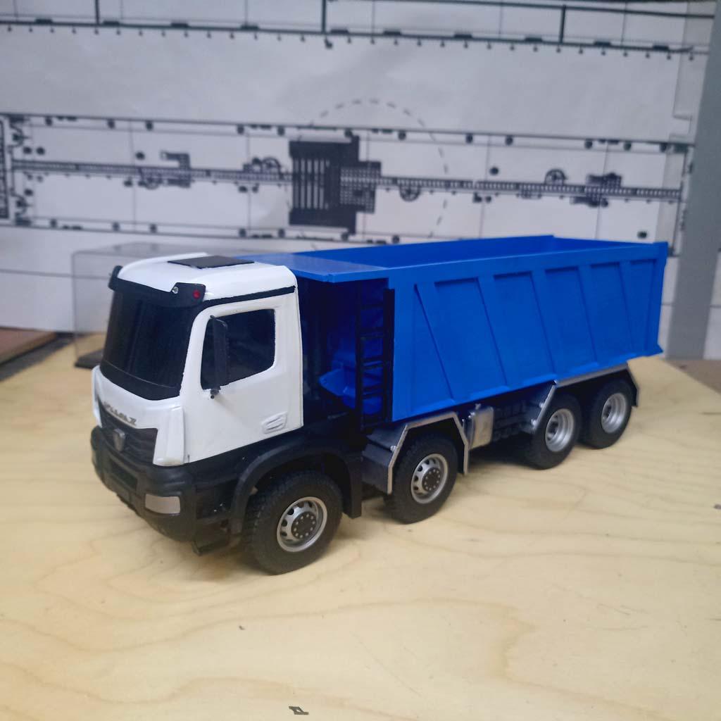 Детально проработанная масштабная модель самосвала КАМАЗ-65951 (масштаб 1:43). Модель демонстрирует четырёхосную конструкцию с массивными колёсами, ярко-синий самосвальный кузов с чёткими рёбрами жёсткости и белую кабину водителя с боковыми зеркалами и дверными ручками. Видны элементы ходовой части, защитные накладки и крепёжные элементы кузова. Реалистичная копия легендарного грузового автомобиля, идеально подходящая для коллекционеров, оформления тематических экспозиций, посвящённых строительной технике, или использования в презентационных материалах производителей спецтехники.