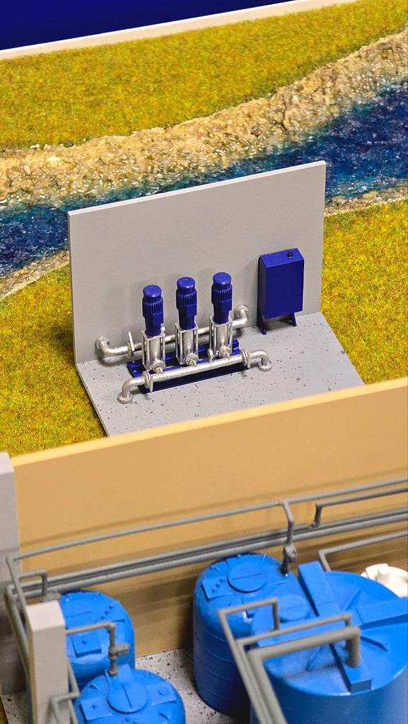 Макет промышленного объекта, насосной станции для перекачки воды с элементами системы водоснабжения или очистки воды. Он демонстрирует работу и взаимодействие технических элементов системы водоснабжения, что важно для проектирования, визуализации и обучения технических специалистов. На переднем плане — несколько больших синих ёмкостей-резервуаров (скорее всего, для хранения воды или химических реагентов). • За резервуарами, ограждённая и вынесенная к реке/каналу, находится площадка, на которой расположены три вертикальных насоса с электрическими приводами и трубопроводной обвязкой. • Также видим шкаф управления (скорее всего, с электроникой для управления насосами). • Все элементы соединены трубопроводами, по которым происходит транспортировка жидкости. • Макет выполнен с отображением окружающей территории — с рекой и травяной зоной.  Для чего это нужно:   • Такая система используется для перекачки воды из природных водоёмов (реки, озёра) на водоочистные или распределительные станции. • Насосы обеспечивают подачу воды в напорные резервуары для дальнейшего распределения по системе водоснабжения (например, в систему питьевой воды города или промышленного предприятия). • Подобные насосные станции также могут использоваться в процессах водоочистки, орошения, промышленного водоснабжения и аварийной подачи воды.