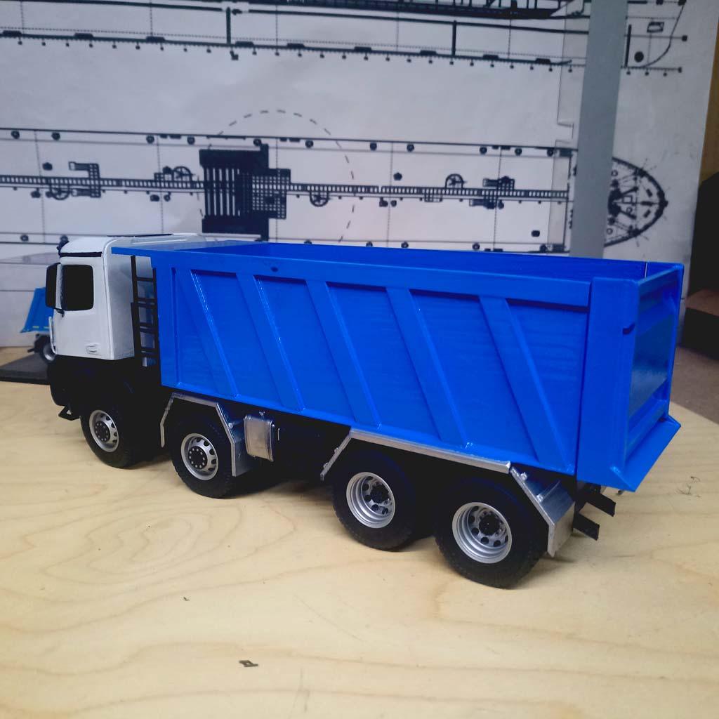 Детально проработанная масштабная модель самосвала КАМАЗ-65951 (масштаб 1:43). Модель демонстрирует четырёхосную конструкцию с массивными колёсами, ярко-синий самосвальный кузов с чёткими рёбрами жёсткости и белую кабину водителя с боковыми зеркалами и дверными ручками. Видны элементы ходовой части, защитные накладки и крепёжные элементы кузова. Реалистичная копия легендарного грузового автомобиля, идеально подходящая для коллекционеров, оформления тематических экспозиций, посвящённых строительной технике, или использования в презентационных материалах производителей спецтехники.