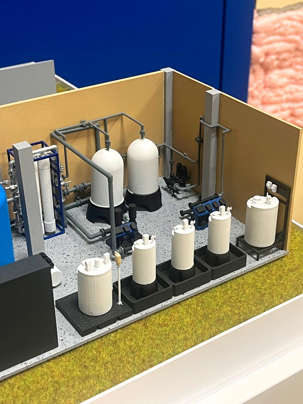 Масштабный макет установки водоочистки или водоподготовки изготовленный для выставки. Такие системы предназначены для очистки воды от примесей, солей, ионов тяжёлых металлов, бактерий и других загрязнений. Обычно используются в промышленности, на предприятиях, в коммунальных хозяйствах, а также для подготовки питьевой воды.  Основные элементы, которые можно выделить на макете: • Большие цилиндрические баки (возможно, фильтры с различными загрузками — угольные, песчаные и другие). • Резервуары/емкости для реагентов или фильтрующих материалов. • Насосы и трубопроводы для перекачки и распределения воды. • Арматура (краны, клапаны) для управления потоками.  Применение таких установок: • Производство питьевой воды • Подготовка воды для котельных и теплоэнергетики • Очистка сточных вод • Водоснабжение промышленных предприятий