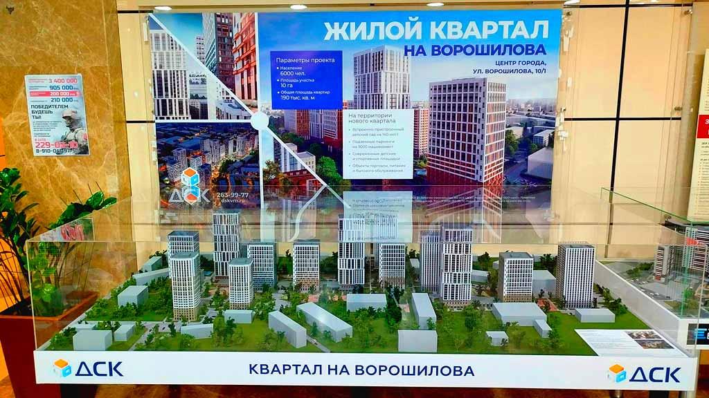 Масштабный 3D-макет жилого квартала, размещенный в стеклянной витрине презентационного стенда. Модель демонстрирует планировочное решение застройки по адресу ул. Ворошилова, 10/1: комплекс современных многоэтажных жилых домов разной высотности с характерной архитектурой фасадов. В макете детально проработана придомовая территория — видны зеленые зоны с деревьями, газоны, пешеходные дорожки и элементы благоустройства. На переднем плане расположена информационная панель с логотипом застройщика «ДСК» и названием проекта «Квартал на Ворошилова». На заднем плане — большой рекламный баннер с визуализацией будущих зданий, параметрами проекта и указанием локации в центре города. Макет служит наглядным инструментом для демонстрации концепции будущей застройки.