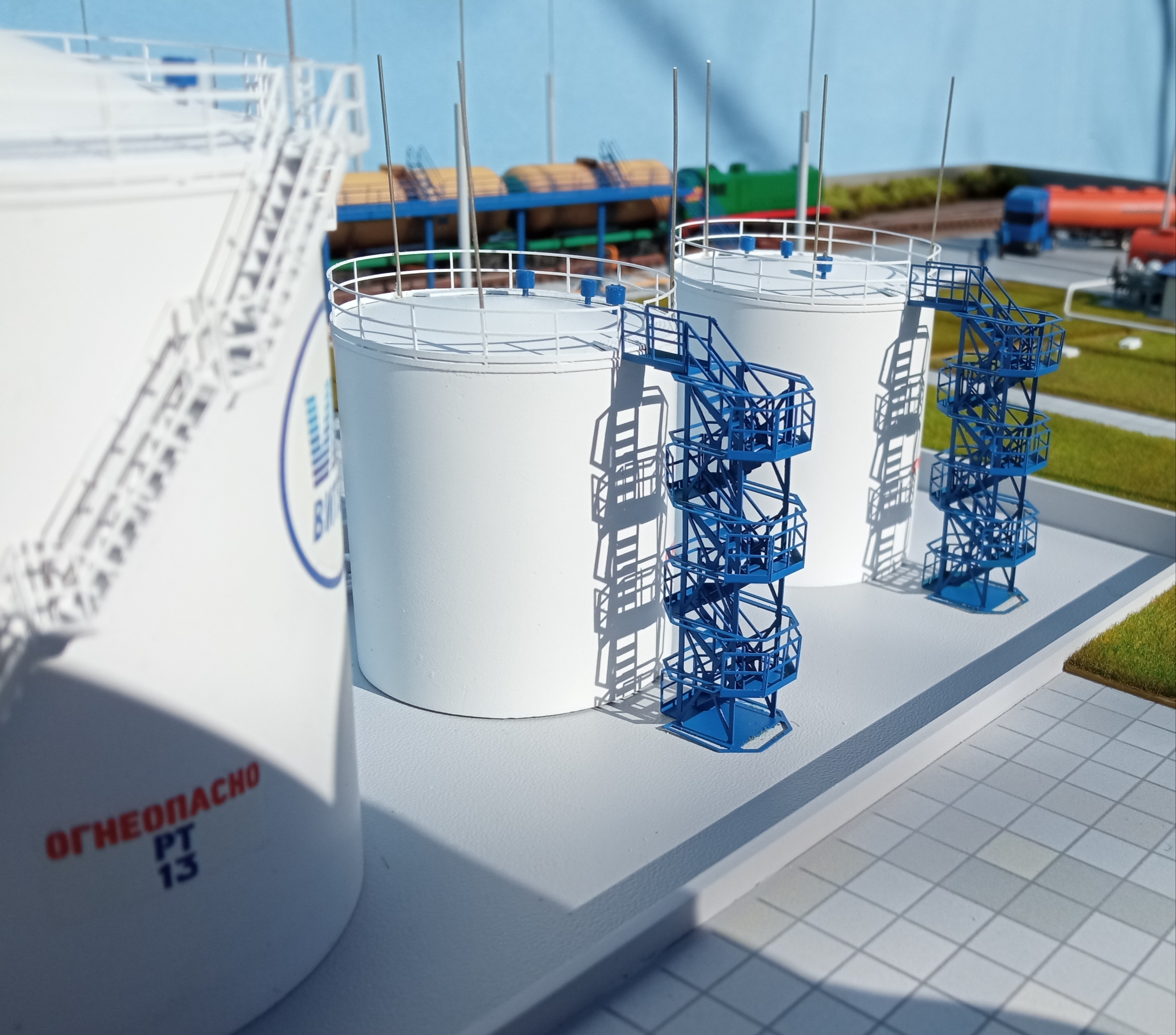 Макет резервуаров нефтебазы. В центре композиции расположены несколько цилиндрических резервуаров, установленных на бетонной площадке. Все резервуары выглядят новыми и ухоженными, без видимых повреждений. Вокруг резервуаров можно заметить подъездные пути и ограждения, обеспечивающие безопасность. Вдалеке виднеются строения, вероятно, административные здания и складские помещения, что указывает на масштабность и значимость нефтебазы.  Эта фотография демонстрирует высокий уровень организации и профессионализма в строительстве и эксплуатации нефтебазы.