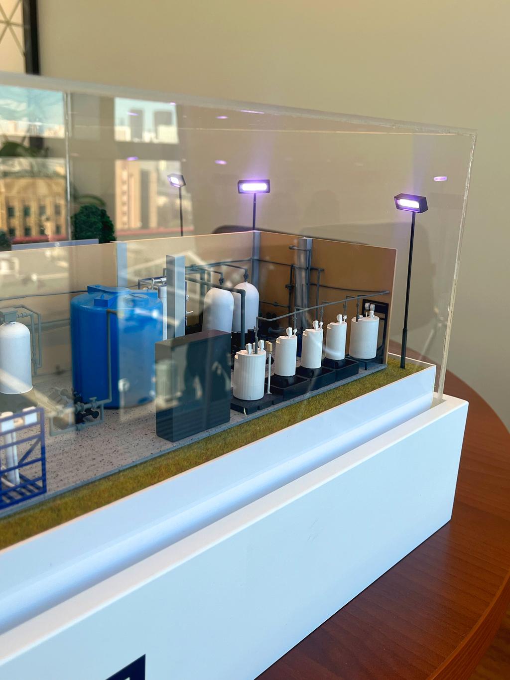 Макет узла очистки и подготовки воды промышленного назначения, изготовленный на заказ в студии макетов для выставки. На макете видны различные резервуары (ёмкости) разного объёма, трубы, а также техническое оборудование. • Большие синие и белые ёмкости имитируют основные элементы системы водоочистки. • Расположены осветительные мачты (уличные фонари), что говорит о внешнем объекте. • Макет защищён прозрачным кожухом.  Макеты такого рода применяются для демонстрации заказчикам, инженерным работникам и для утверждения проектов на стадиях проектирования и строительства.