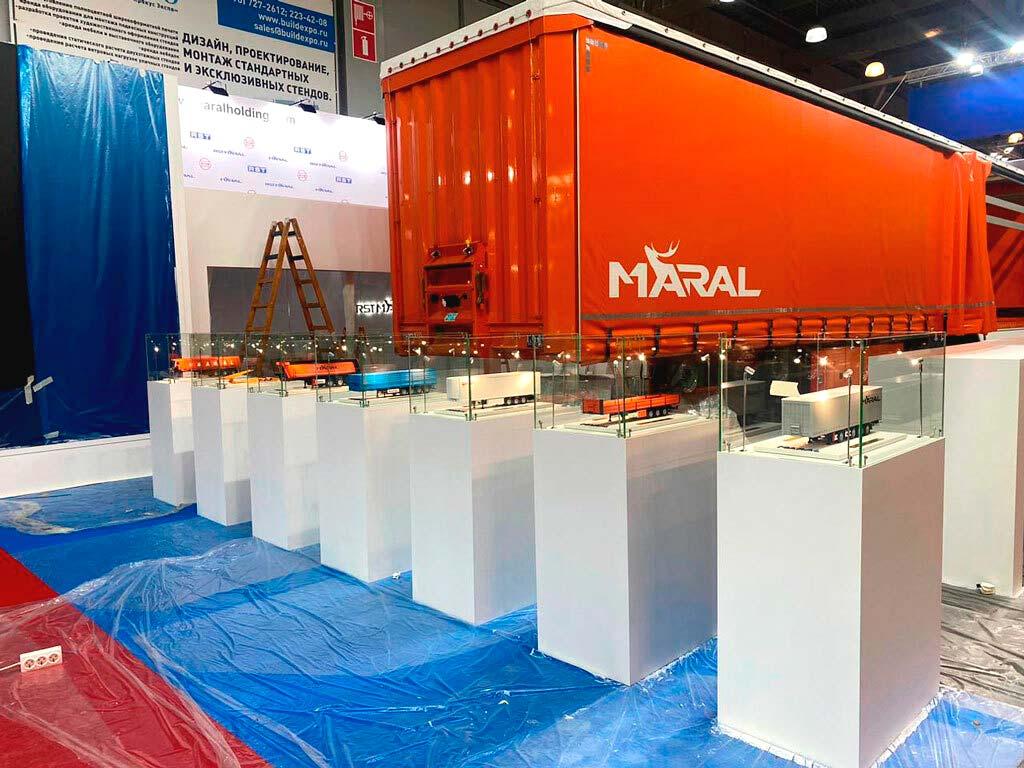 На фотографии представлен макет оранжевого полуприцепа с 12 колесами, выполненный с высокой детализацией. Полуприцеп имеет аккуратные линии и реалистичную отделку, что подчеркивает его предназначение для выставки. Фон изображения ненавязчивый, что позволяет сосредоточиться на модели техники.