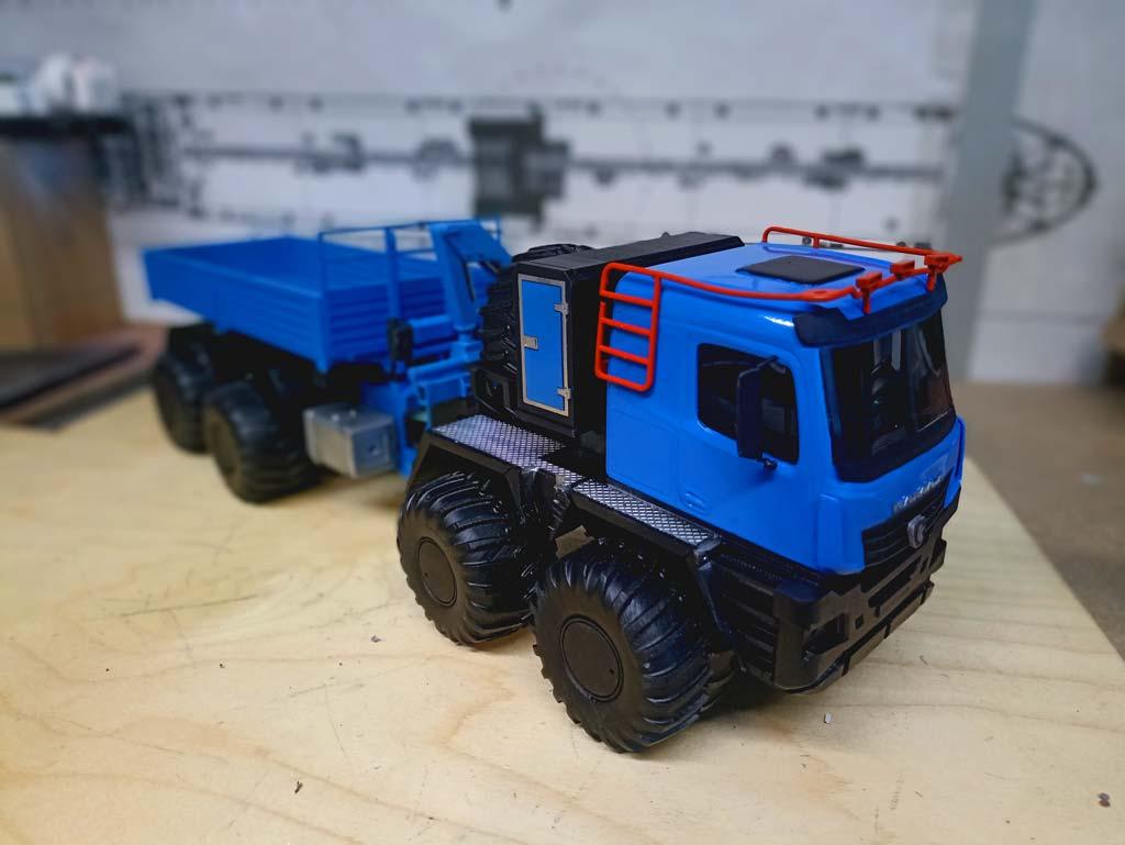 Детализированная масштабная модель грузового автомобиля КАМАЗ с прицепом (масштаб 1:43). Кабина выполнена в насыщенном синем цвете с контрастными чёрными элементами, дополнена красными рейлингами на крыше и боковыми лестницами. Массивные чёрные колёса с выраженным протектором подчёркивают внедорожный характер модели. Прицеп также окрашен в синий цвет, оснащён бортами и реалистичными крепёжными элементами. Модель демонстрирует ключевые детали: решётку радиатора с логотипом КАМАЗ, боковые зеркала, дверные ручки, элементы ходовой части. Идеально подходит для коллекционирования, оформления выставок, посвящённых грузовой технике, или использования в презентационных материалах производителей автотранспорта.