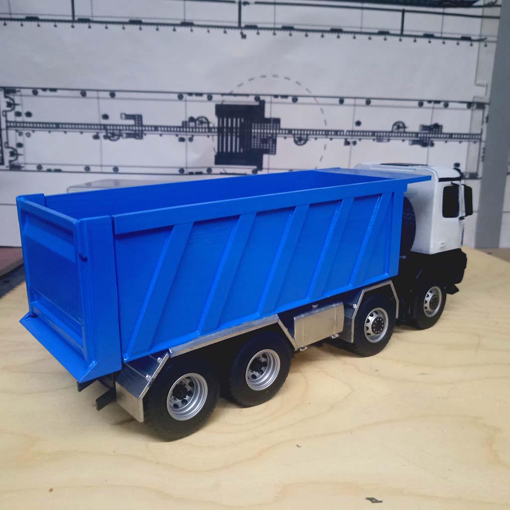 Детально проработанная масштабная модель самосвала КАМАЗ-65951 (масштаб 1:43). Модель демонстрирует четырёхосную конструкцию с массивными колёсами, ярко-синий самосвальный кузов с чёткими рёбрами жёсткости и белую кабину водителя с боковыми зеркалами и дверными ручками. Видны элементы ходовой части, защитные накладки и крепёжные элементы кузова. Реалистичная копия легендарного грузового автомобиля, идеально подходящая для коллекционеров, оформления тематических экспозиций, посвящённых строительной технике, или использования в презентационных материалах производителей спецтехники.