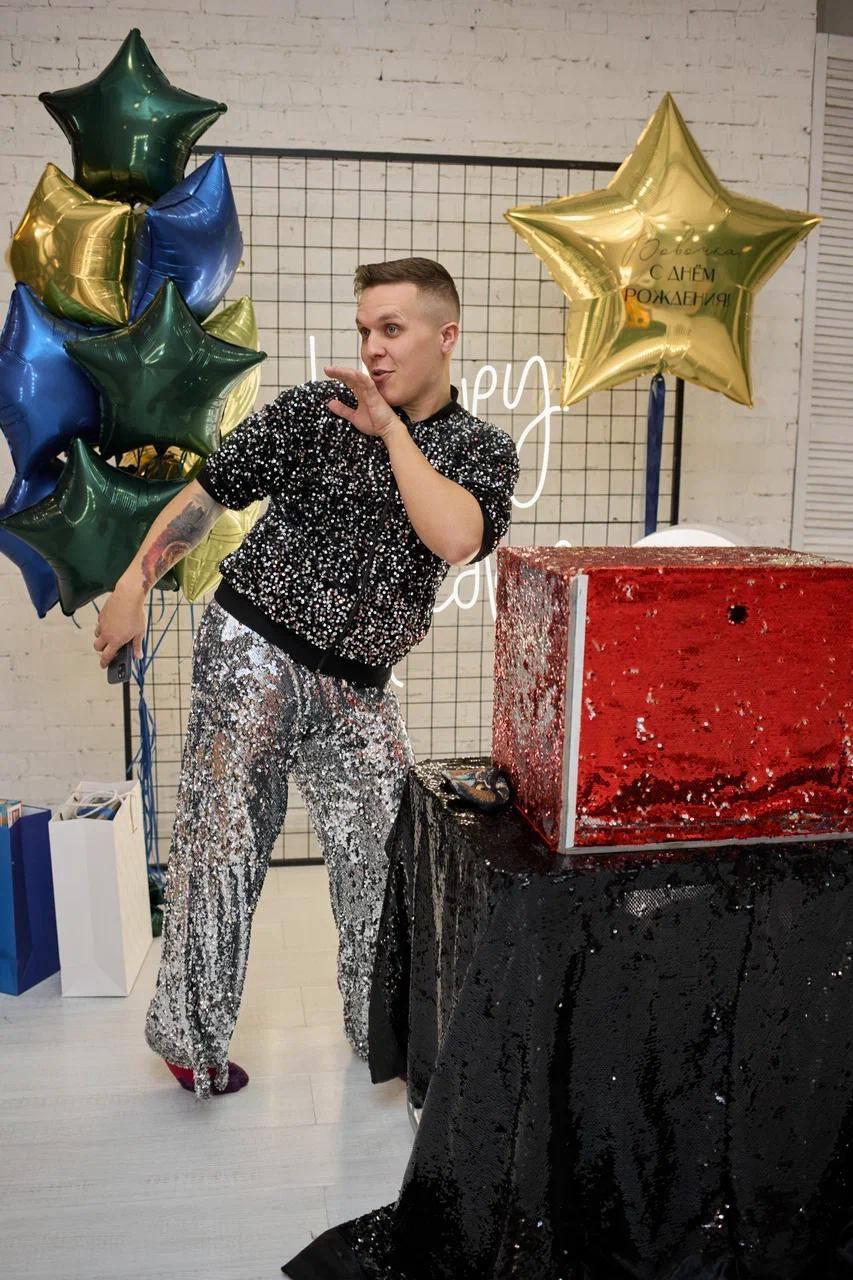 Ведущий программы Треш-коробка от AMStudio развлекает гостей на празднике, рядом яркая коробка с сюрпризом и праздничный декор с шарами